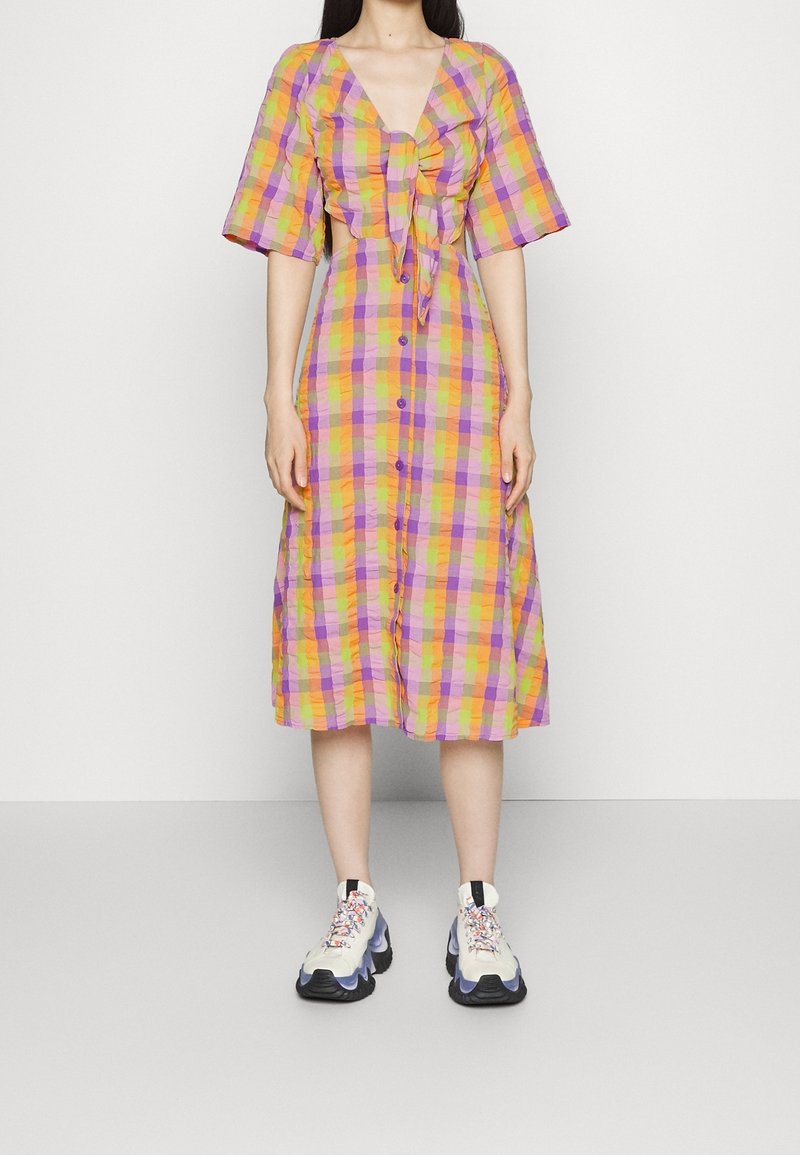 Robe à carreaux jaune, violette et orange, avec un col en V, des manches courtes, un nœud à l'avant et une fermeture boutonnée. Associée à des baskets épaisses.