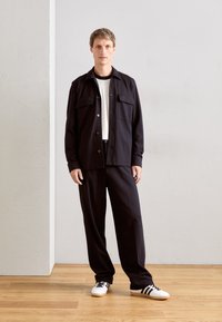 Uomo con giacca nera e pantaloni, camicia bianca e sneakers bianche con strisce nere, in piedi su un pavimento di legno davanti a una parete semplice.