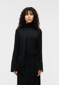 Μαύρο φόρεμα με μακριά μανίκια, ψηλό λαιμό και λεπτομέρεια με ρέον φουλάρι. Κατασκευασμένο από λείο ύφασμα, διαθέτει φαρδιά μανίκια και διακριτική υφή.