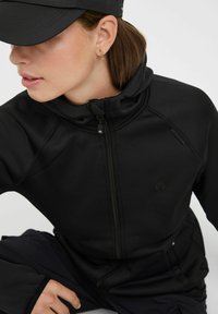 Zwarte zip-up jas met hoge kraag en aangesloten ontwerp. Zacht, rekbaar materiaal met subtiele logo-details. Gecombineerd met een bijpassende zwarte onderkant.