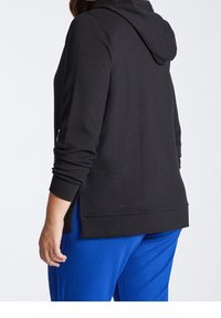 Černá žebrovaná mikina s texturovaným designem, dlouhými rukávy a bočními rozparky, spárovaná s modrými tepláky. Tkanina vypadá měkká a elastická.