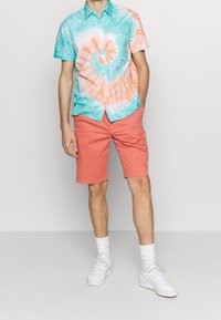 Maglietta a maniche corte in tie-dye turchese e corallo con un motivo a spirale, abbinata a pantaloni corti rosa. Sneakers bianche e calzini completano l'outfit.