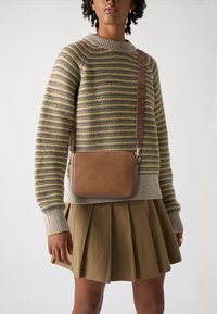 Borsa a tracolla in pelle marrone con logo impresso, chiusura a zip e tracolla regolabile. Indossata sopra un maglione lavorato a righe e una gonna plissettata.