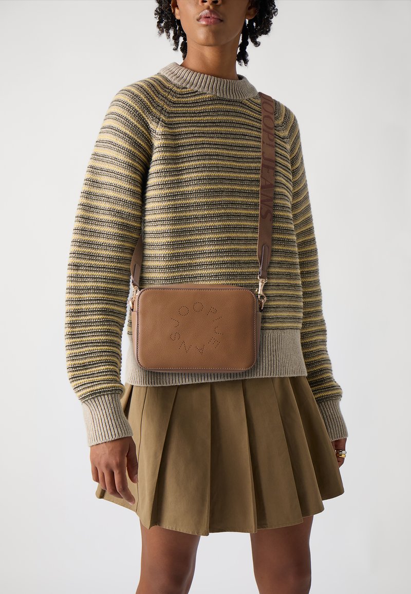 Borsa a tracolla in pelle marrone con logo impresso, chiusura a zip e tracolla regolabile. Indossata sopra un maglione lavorato a righe e una gonna plissettata.