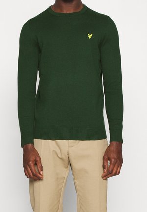 Pull à manches longues vert foncé avec un petit logo d'oiseau jaune sur la poitrine, porté avec un pantalon beige.