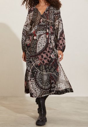 Femme portant une longue robe à motifs mixtes floraux et géométriques avec des bottes noires épaisses, debout à l'intérieur contre un mur uni.
