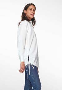 Weißes Baumwollhemd mit langen Ärmeln und hohem Kragen, seitlichen Bindebändern und einer leicht lockeren Passform, kombiniert mit dunklen Jeans.