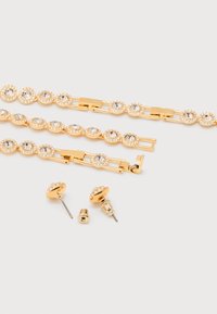 Σετ βραχιολιού σε χρυσό τόνο και αντίστοιχα σκουλαρίκια καρφάκια, κάθε κομμάτι διακοσμημένο με καθαρά στρογγυλά κρύσταλλα σε κυκλικό πλαίσιο σε λευκό φόντο.