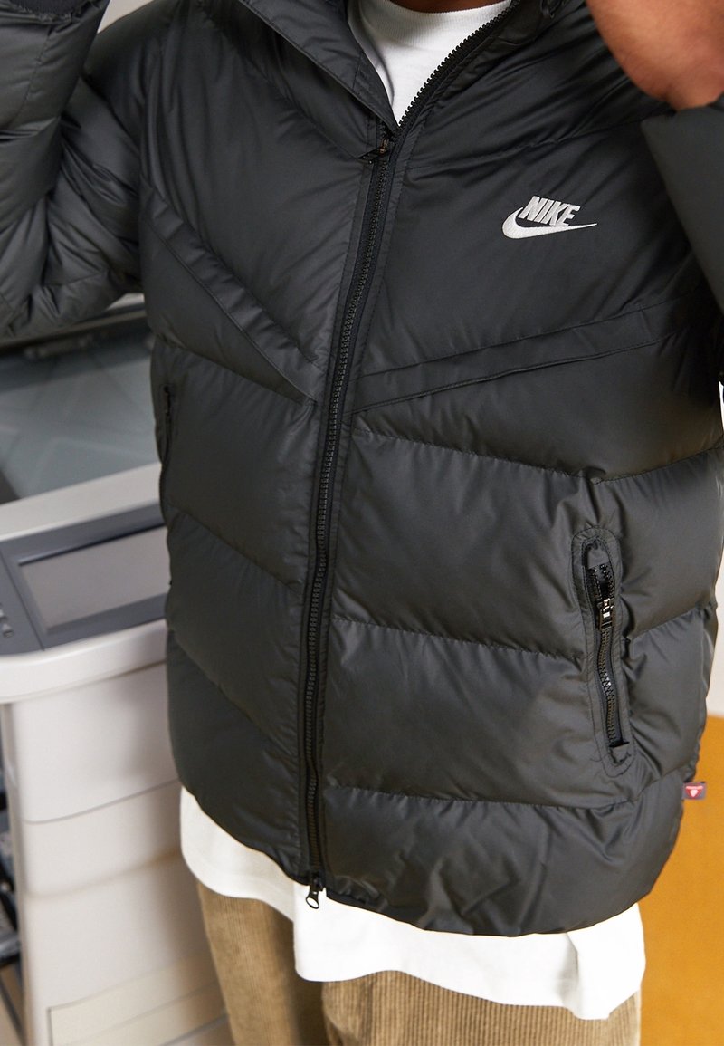 Black puffer jacket with a zip closure, featuring a Nike logo on the chest, side pockets, and horizontal quilted design.