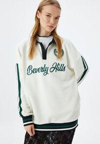 Felpa bianca con colletto nero, dettagli verde e testo "Beverly Hills" in corsivo; presenta maniche a righe e una finitura testurizzata.