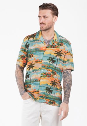 Homme avec des tatouages portant une chemise hawaïenne colorée à manches courtes avec des palmiers, des fleurs et une scène de plage, associée à un pantalon blanc.