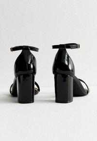 Czarne sandały na obcasie z lakierowanej skóry, z tylnym paskiem na kostkę, masywnym obcasem i otwartym noskiem. Minimalistyczny styl o gładkiej teksturze.
