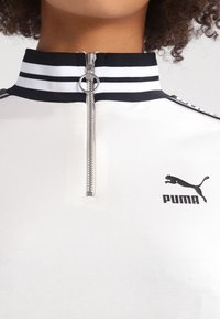 Bílá zipová mikina Puma s černým a bílým pruhovaným límcem a stříbrným zipem, kterou nosí osoba s kudrnatými vlasy.