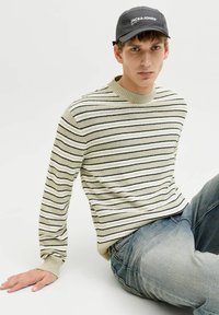 Jeune homme portant une casquette noire, un pull à rayures beige et noir, et un jean bleu, assis avec une main posée sur le sol, devant un fond uni.