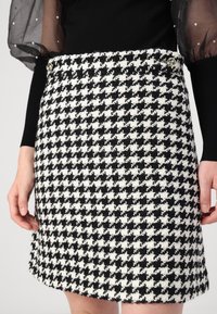 Svart och vit hundtandsmönstrad kjol i texturerad tyg, med en figursydd silhuett och metalliska detaljer vid midjan, visad mot en neutral bakgrund.