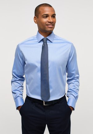 Hellsblaues Langarmhemd mit spitzem Kragen, kombiniert mit einer strukturierten dunkelblauen Krawatte und einer Marineblauen Hose, die Taschen und einen Gürtel aufweist.