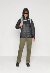 Μαύρο μπουφάν puffer με κουκούλα, φερμουάρ μπροστά, καπιτονέ υφή· παντελόνι ελιάς πράσινο· μαύρα παπούτσια· σιέλ σακίδιο πλάτης· σκούρα γυαλιά ηλίου.