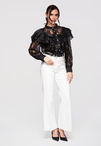 Femme debout, main sur la hanche, portant un chemisier en dentelle noire transparente avec des volants, un pantalon blanc taille haute, une ceinture noire et des talons noirs, sur fond uni.