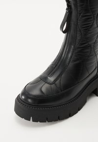 Bottine matelassée noire avec une tige en cuir brillant, un motif texturé, une fermeture éclair sur le côté et une semelle en caoutchouc épaisse avec des détails striés.