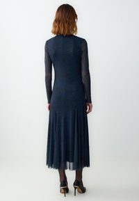 Abito lungo navy con maniche lunghe, busto aderente e orlo fluido; sovrapposizione trasparente con una texture sottile; abbinato a scarpe con tacco alto nere.