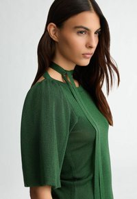 Blusa verde con maniche corte, texture lucida, spalle scoperte e design a colletto all'americana con lunghi lacci lungo la parte anteriore.