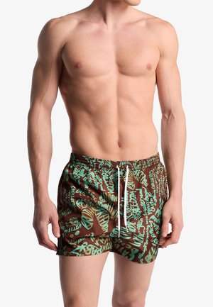 Uomo senza maglietta che indossa pantaloncini da bagno marroni con testo verde in stile graffiti e coulisse bianca, in piedi su sfondo bianco.