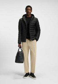 Giacca a vento nera con cappuccio, maglione a maglia testurizzato, pantaloni beige, sneakers nere e borsa in pelle. Materiali lisci e strutturati.