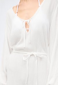 Λευκό φόρεμα με υφή από ύφασμα, μακριά μανίκια, ντεκολτέ V με δέσιμο, ζώνη στη μέση και ορατές ροζ τιράντες σουτιέν σε άτομο.