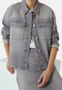 Graue Jeansjacke mit zwei Fronttaschen, Knopfverschluss und umgeschlagenen Ärmeln. Glatte Textur und dezente Farbverläufe.