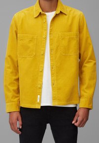 Chaqueta camisa de pana amarilla con cierre de botones, dos bolsillos en el pecho y una camiseta blanca debajo, combinada con pantalones negros.