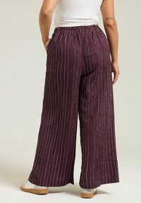 Un pantalon large en tissu bordeaux texturé avec des rayures blanches verticales. Taille élastique avec une coupe décontractée, associé à des baskets blanches.