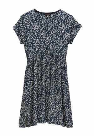 Kvinde iført en blå blomsterkortærmet kjole og brune cowboystøvler, der står på en jævn gulv mod en beige baggrund.
