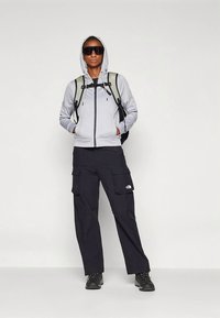 Γκρι φούτερ με φερμουάρ και κουκούλα, συνδυασμένο με μαύρο παντελόνι cargo με μεγάλες τσέπες και μαύρες μπότες. Ορατό σακίδιο με ιμάντες.