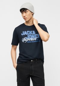 Νέος άνδρας που φοράει γκρι πλεκτό σκούφο, μπλε σκούρο γραφικό μπλουζάκι με το κείμενο "Jack & Jones Superior" και μαύρο παντελόνι cargo, ποζάροντας με το ένα χέρι στον λαιμό.