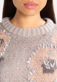 Maglione a maglia grigio chiaro con colletto a coste, caratterizzato da design astratti in pesca e marrone e cuciture decorative in fili di contrasto.