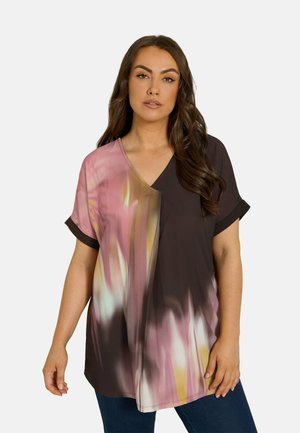 Vrouw draagt een losse V-hals top met korte mouwen met een abstract patroon in roze, bruin en geel en een donkerblauwe broek, staand tegen een effen achtergrond.