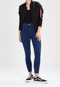 Chaqueta bomber negra con puños acanalados, vaqueros ajustados de tiro alto en azul oscuro y zapatillas Vans negras con suela blanca y detalles de cordones.
