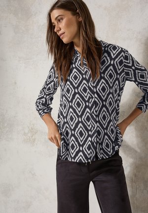 Camicia a bottone con un motivo a rombi blu e bianchi, maniche lunghe e vestibilità comoda, abbinata a pantaloni scuri con una texture liscia.