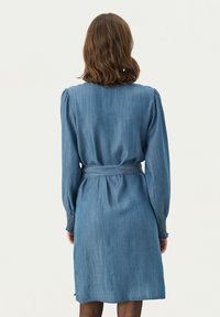 Robe en denim avec une taille cintrée, des manches longues et un détail froncé aux poignets. De couleur bleu moyen avec un ourlet à mi-mollet.