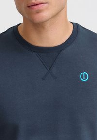 Granatowy sweatshirt z ribbowym kołnierzem i niebieskim haftowanym logo. Tkanina wydaje się miękka, ma gładką teksturę i widoczne detale szycia.