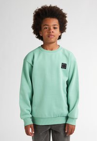 Sweat-shirt vert menthe avec des poignets et un ourlet côtelés. Présente un patch noir carré avec un motif circulaire à l'avant. Texture douce.
