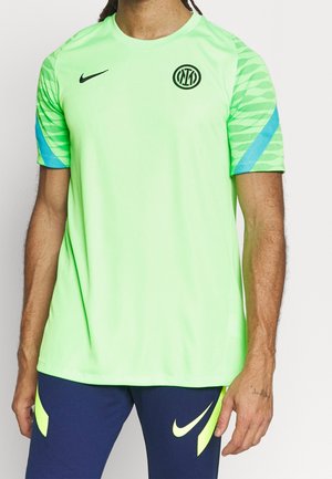 Man draagt een neongroen sportshirt met korte mouwen, met een zwart Inter Milan-logo en Nike-swoosh, gecombineerd met marineblauwe broek met neonaccenten.