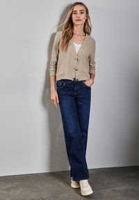 Beige cardigan med knappar, bärs över en vit topp; ihop med mörkblå jeans och beige sneakers mot en grå bakgrund.