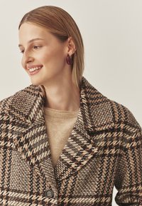 Bruin en beige houndstooth-patroon jas met een grote kraag, knoopsluiting, gedragen over een beige trui, gecombineerd met druppelvormige oorbellen.