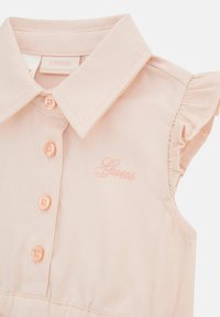 Abito rosa chiaro in cotone, con colletto, maniche a volant e cinque bottoni. Include il logo ricamato "Guess" nello stesso colore.