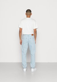 Jeans de ganga azul claro com um corte relaxado, bolsos traseiros e um patch de couro bege na cintura; combinados com uma t-shirt branca simples.