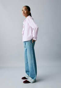 Cămașă roz cu dungi, cu detalii de volă, blugi largi albaștri cu tivuri răsucite și pantofi slip-on vișinii. Fundal gri simplu.