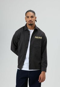 Zwart geruit overhemd met een kraag, knoopsluiting, en "FREE DOM" geborduurd in geel op de linkerborst. Draagt over een wit shirt.