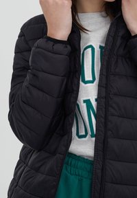 Personne portant une veste matelassée noire par-dessus un sweat-shirt gris avec des lettres vertes, ajustant le col avec les deux mains.
