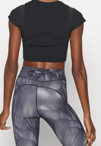 Černý krátký top s texturovanými, třpytivými rukávy spárovaný s šedými legínami s geometrickým vzorem a kontrastním prošíváním.